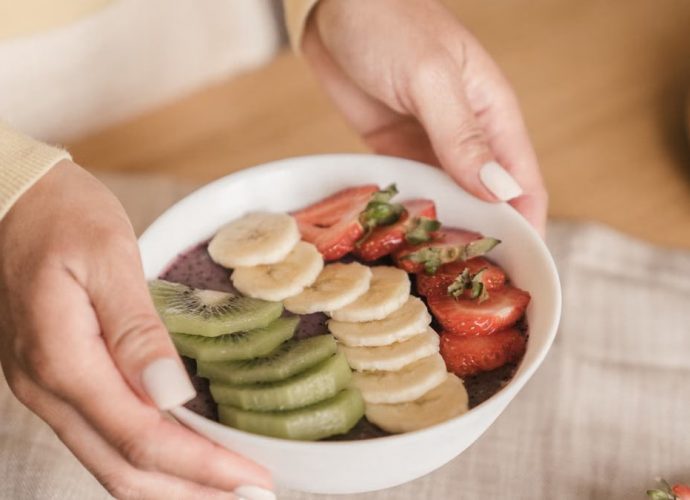 Delicious smoothie bowl topped with fresh fruits like kiwi, banana, and strawberries, held by manicured hands.