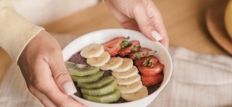 Delicious smoothie bowl topped with fresh fruits like kiwi, banana, and strawberries, held by manicured hands.