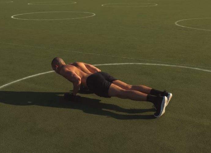A shirtless man performing push-ups on a green field under bright daylight.