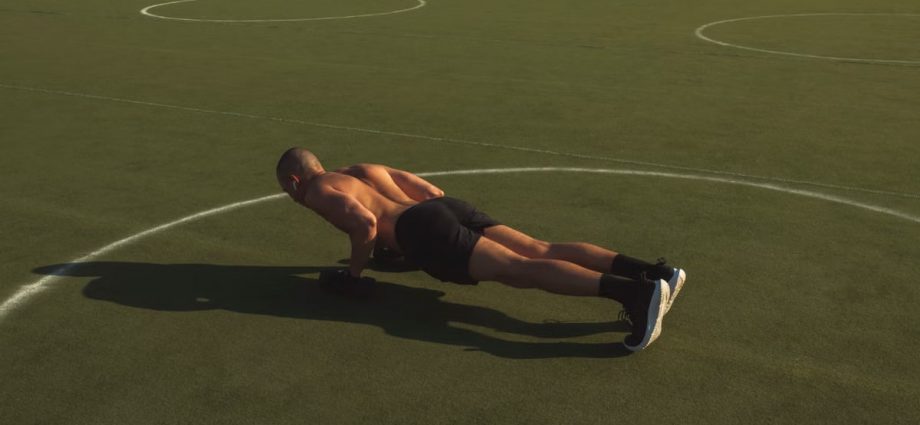 A shirtless man performing push-ups on a green field under bright daylight.